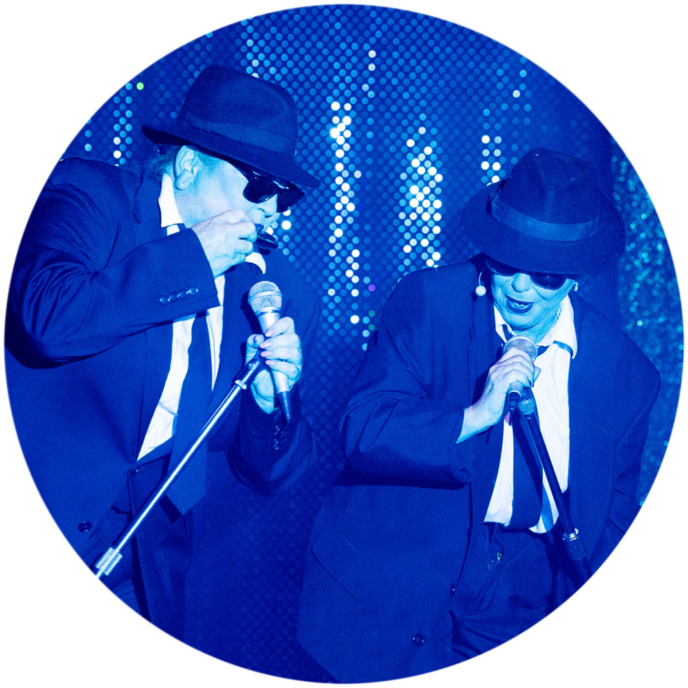 Eine Szene aus dem Musik-Kabarett Stars on Stage zeigt zwei Darstellerinnen, die ein Lied aus dem Film The Blues Brothers nachstellen. Die Bühne ist in blaues Licht getaucht, was eine coole, mysteriöse Atmosphäre schafft. Die Darstellerinnen tragen die ikonischen Kostüme der Blues Brothers: schwarze Anzüge, weiße Hemden, schwarze Hüte und Sonnenbrillen. Sie stehen mit coolen Posen und bewegen sich synchron, während sie das Lied mit voller Energie und viel Leidenschaft performen.
