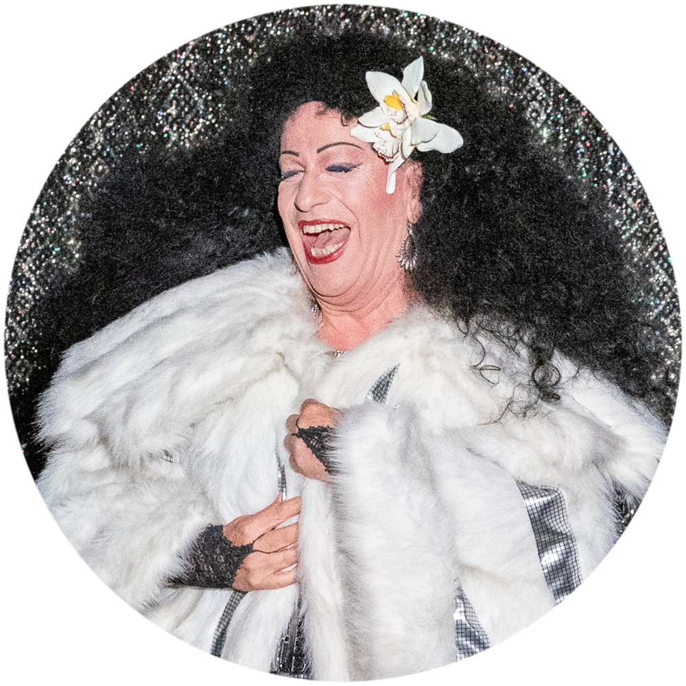 Eine Szene aus dem Musik-Kabarett Stars on Stage zeigt einen Mann, der das berühmte Lied Chain Reaction von Diana Ross mimt. Er trägt einen glänzenden weißen Perlzmantel und eine auffällige, schwarz stark gelockte Perücke, die an Diana Ross’ ikonischen Look erinnert. Mit viel Energie und Leidenschaft performt er das Lied, während er die markanten Bewegungen und Ausdrucksformen von Diana Ross nachahmt, was die Darbietung zu einem glamourösen Höhepunkt der Show macht.