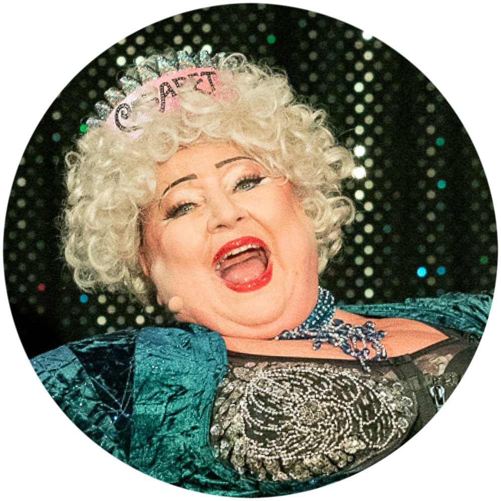 Eine Szene aus dem Musik-Kabarett Stars on Stage zeigt eine blonde Dame mit einer gelockten Perücke, die ein Lied auf der Bühne singt. Sie trägt eine auffällige Krone, auf der das Wort Caberet prangt, was ihr eine majestätische Ausstrahlung verleiht. Ihre Darbietung ist charmant und voller Energie, wobei sie das Publikum mit ihrer Präsenz fesselt. Das Lied, das sie singt, ist nicht bekannt, doch ihre Ausstrahlung macht sie zu einem echten Highlight der Show.