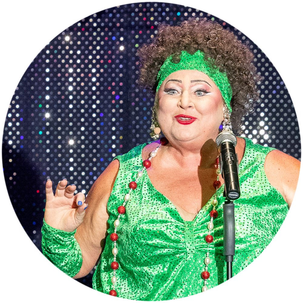 Eine Szene aus dem Musik-Kabarett Stars on Stage zeigt eine Dame, die als Trude Herr verkleidet ist. Sie trägt ein auffälliges grünes Kleid und steht mit lebhafter Mimik auf der Bühne, während sie das bekannte Lied Ich will keine Schokolade performt. Ihre Darstellung ist humorvoll und voller Energie.