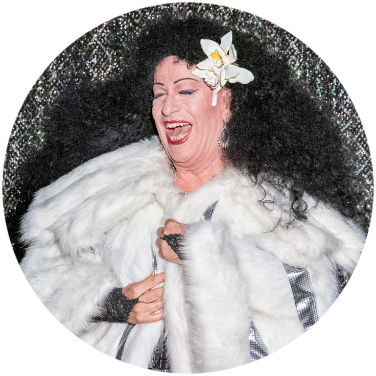 Eine Szene aus dem Musik-Kabarett Stars on Stage zeigt einen Mann, der das berühmte Lied Chain Reaction von Diana Ross mimt. Er trägt einen glänzenden weißen Perlzmantel und eine auffällige, schwarz stark gelockte Perücke, die an Diana Ross’ ikonischen Look erinnert. Mit viel Energie und Leidenschaft performt er das Lied, während er die markanten Bewegungen und Ausdrucksformen von Diana Ross nachahmt, was die Darbietung zu einem glamourösen Höhepunkt der Show macht.